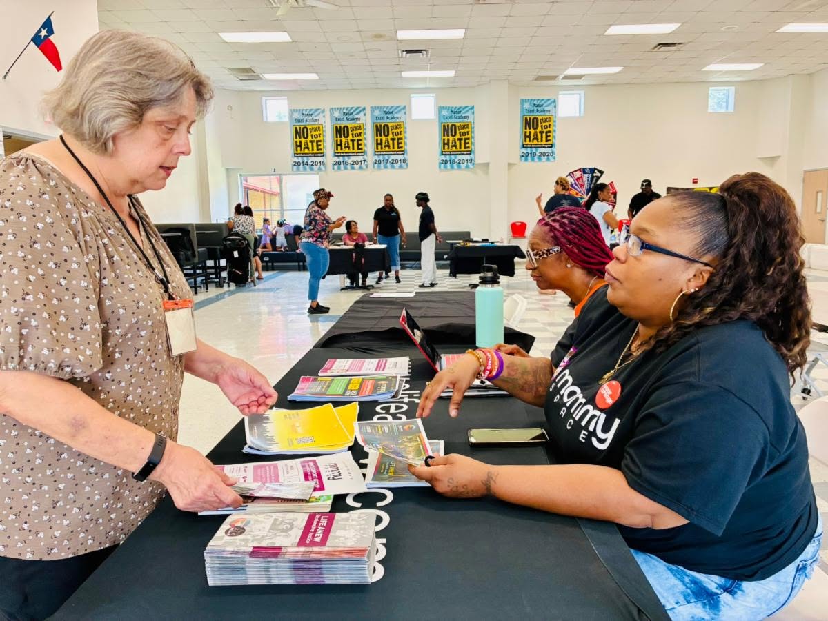 A woman reviews community outreach materials with two staff members at a table during an indoor event, with “No Place for Hate” banners hanging in the background.