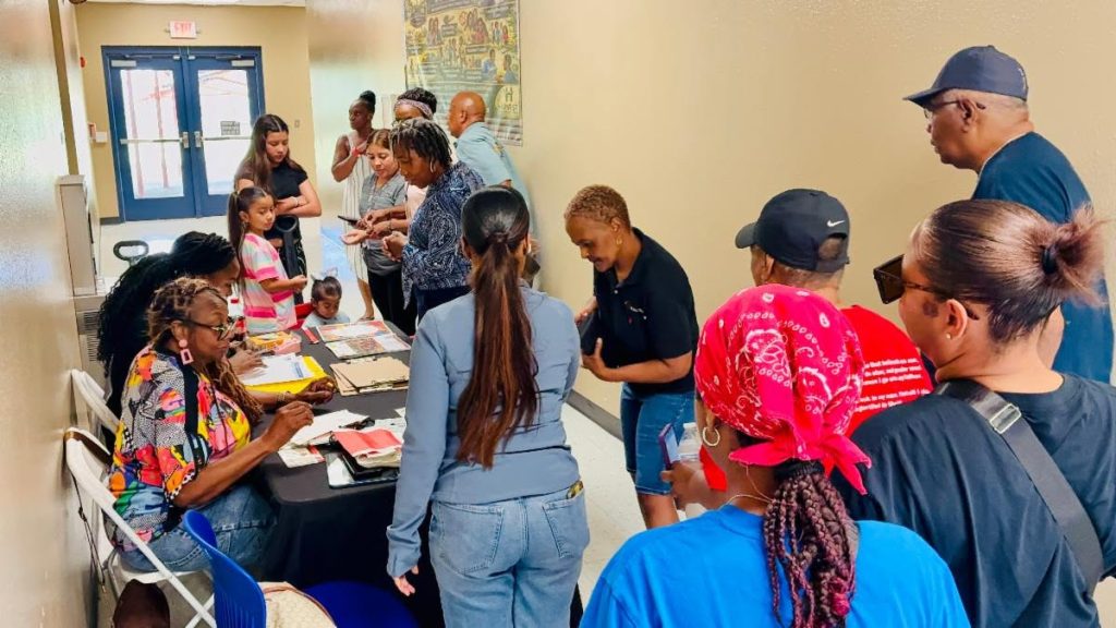 A busy community event check-in area shows adults and children standing in line while volunteers assist at a table covered with papers and supplies.