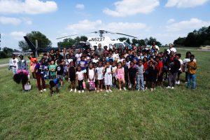 A large group of children and adults pose in a grassy field in front of a white helicopter under a partly cloudy sky.