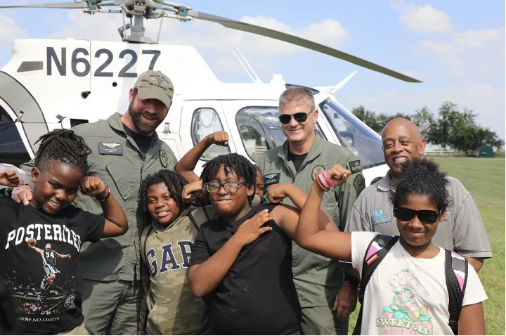 A small group of children and AAYHF CEO Michael Lofton pose with two APD Air Operations Officers in a grassy field in front of a Austin PD helicopter.